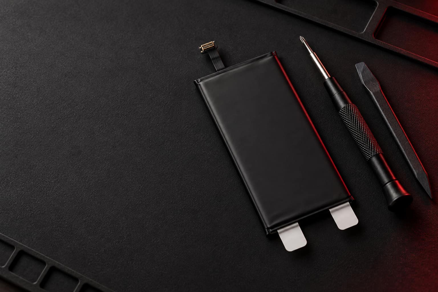 Batería de repuesto nueva y plana, con su conector flex y sus lengüetas, sobre un tapete de reparación junto a un destornillador de precisión y una espátula