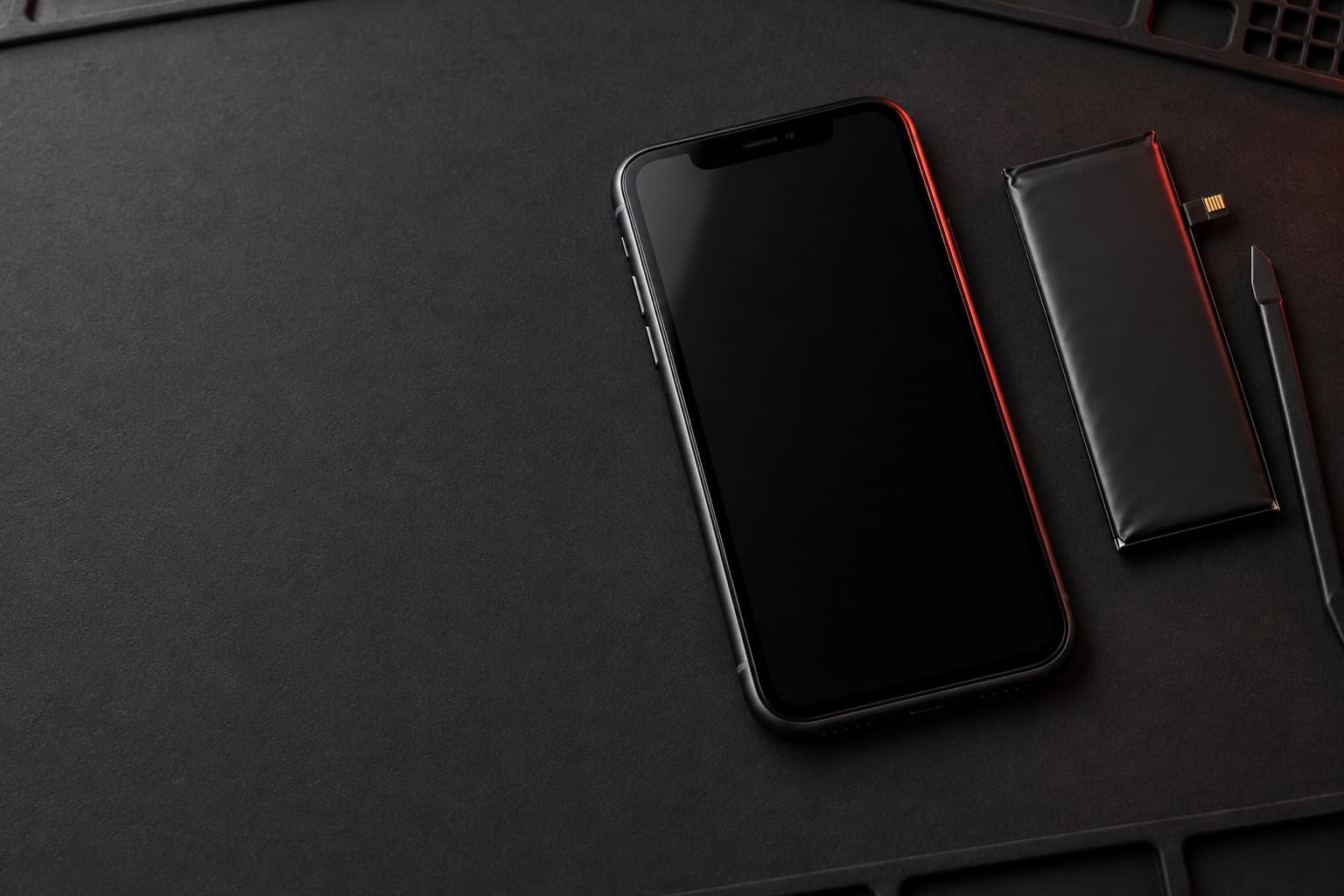 Black iPhone 11 lying face-up on a repair mat beside a replacement battery and a plastic spudger