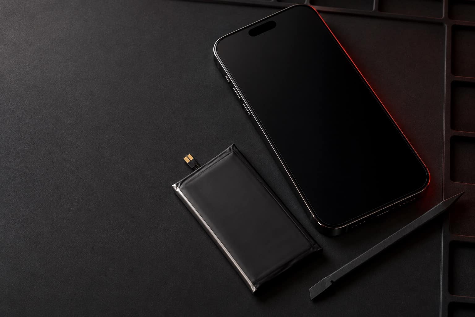 iPhone 14 Pro Max negro espacial sobre un tapete de reparación, junto a una batería de repuesto y una espátula